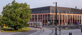 Raitiovaunu ohittaa modernin tiilirakennuksen, jossa on mustia yksityiskohtia ja suuret ikkunat. Lähellä on suuri puu ja useita polkupyöriä.